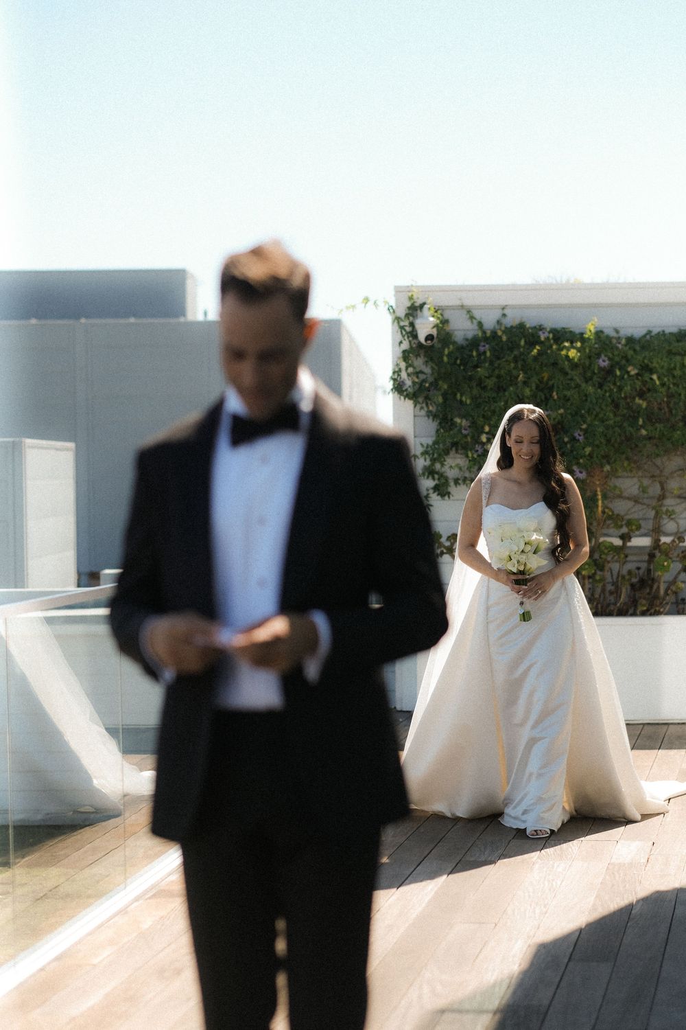 Bride in white gown holding bouquet smiles at groom in black tuxedo on a sunlit rooftop terrace.