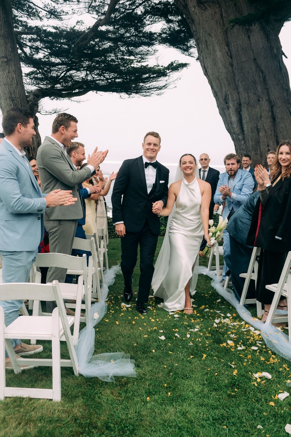 Newlywed couple walks down outdoor wedding aisle lined with white chairs and cheering guests beneath tall trees.