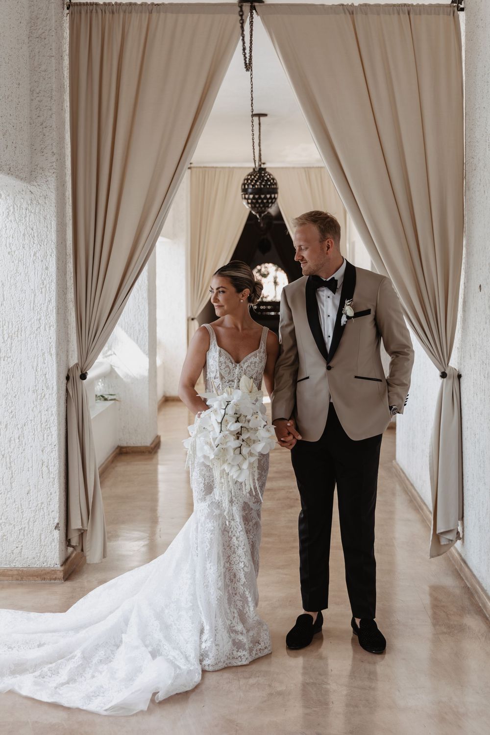 A bride in a flowing white lace gown walks down an elegant hallway with a companion in formal attire.