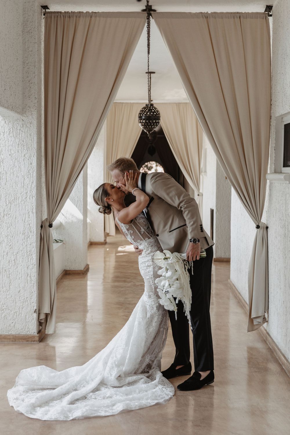 A romantic wedding couple shares intimate kisses in a light-filled hallway with elegant cream curtains and hardwood floors.