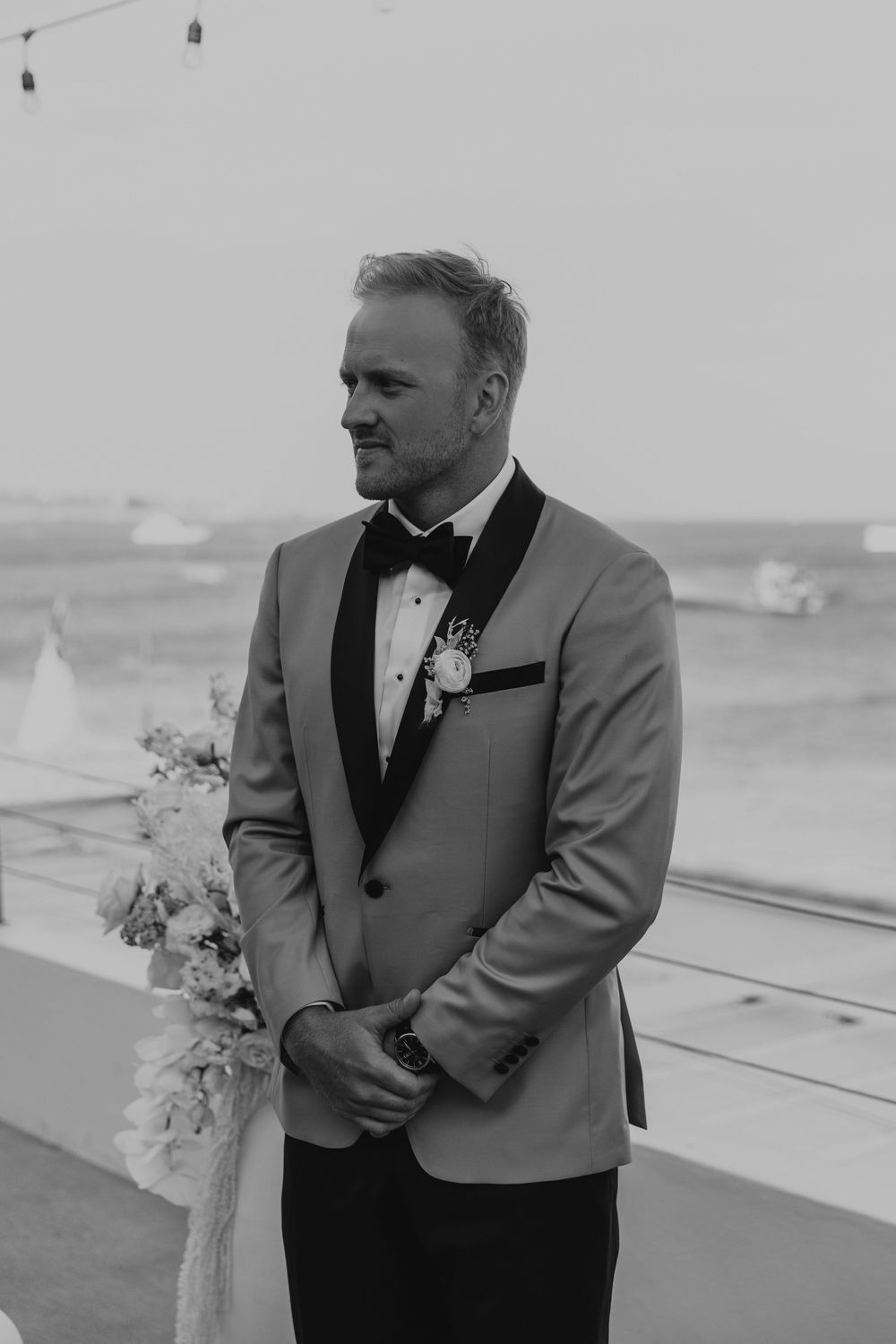 A groom in a gray tuxedo jacket with black lapels stands at an outdoor venue overlooking sailboats on the water.