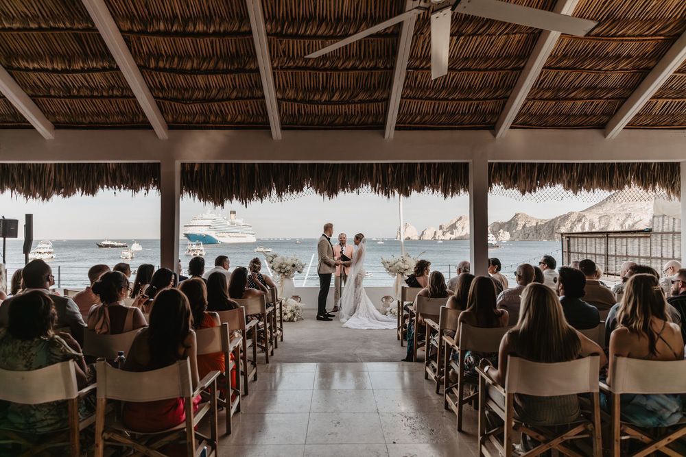 Wedding ceremony guests seated in rows under a thatched roof palapa overlooking the ocean at a tropical beach venue.