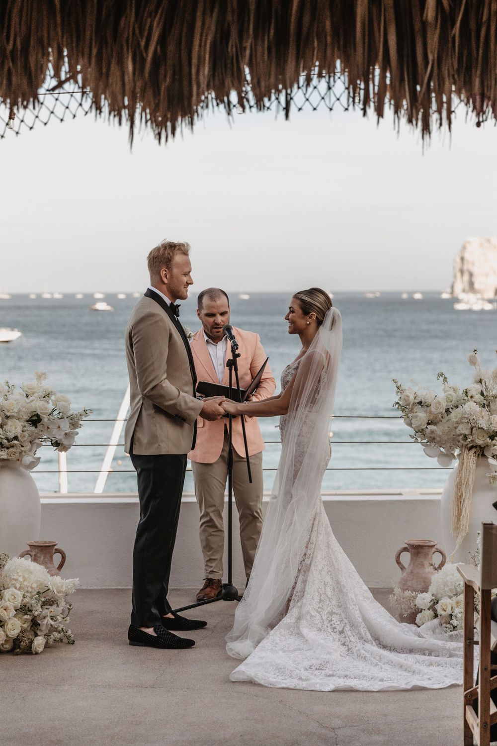 A wedding ceremony takes place on a waterfront terrace with palm fronds overhead and white floral decorations.