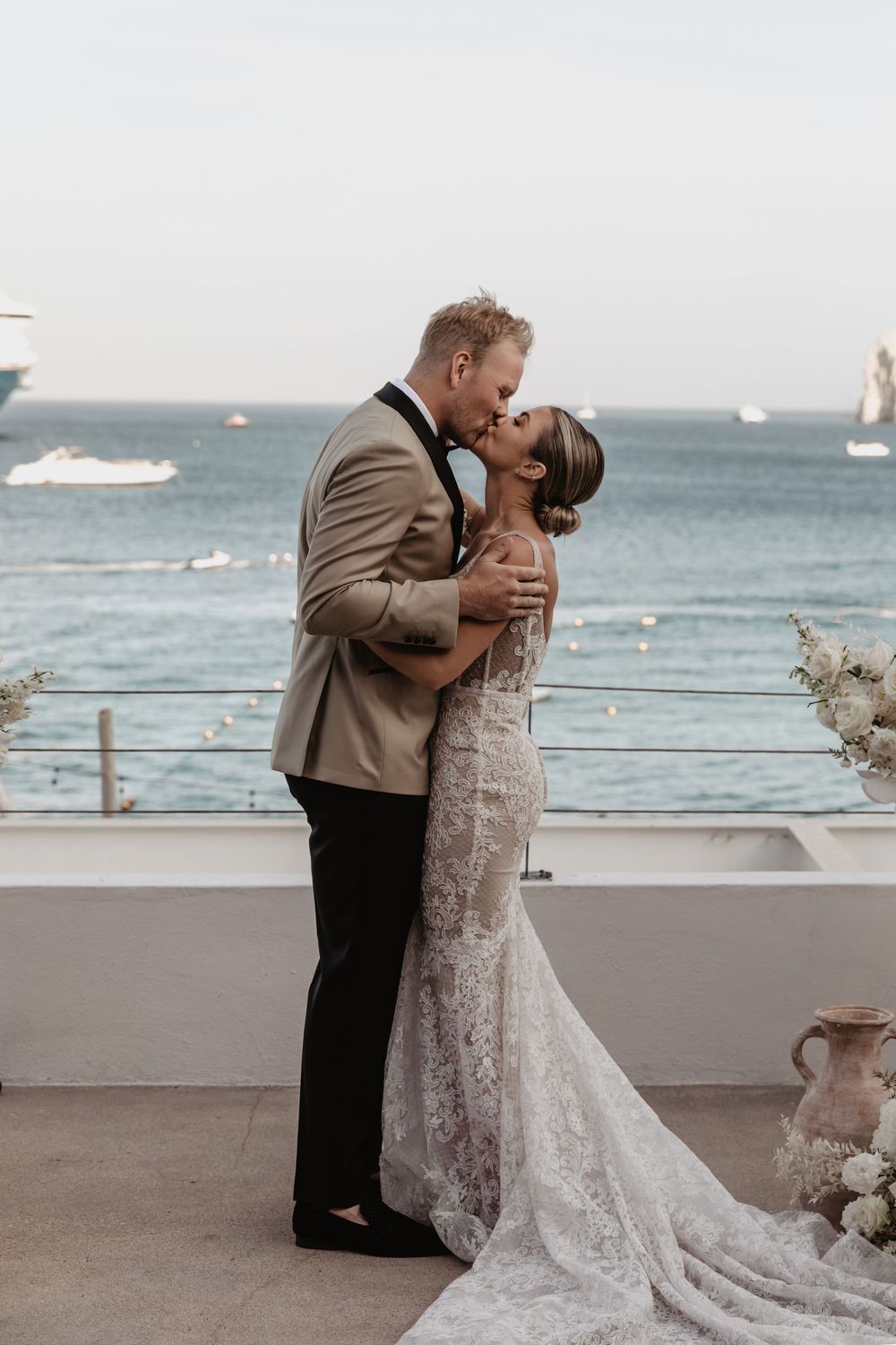A couple shares a romantic kiss on an oceanfront terrace decorated with flowers, with sailboats visible in the background.