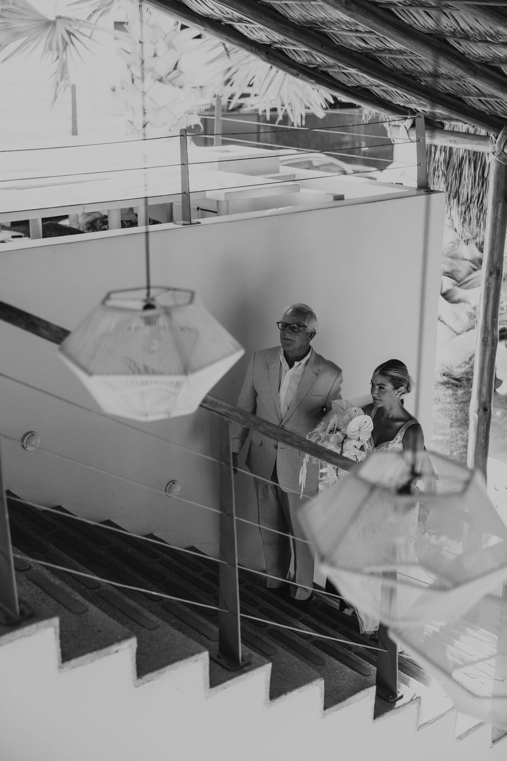 A black and white photo of wedding guests descending a staircase under hanging geometric light fixtures.