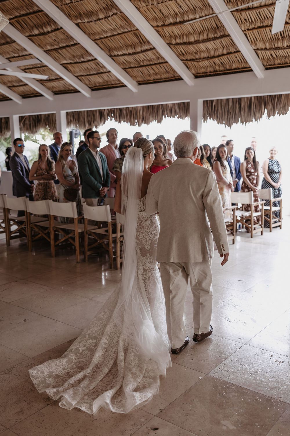 A destination beach wedding ceremony takes place under a thatched-roof palapa with guests seated in white chairs.