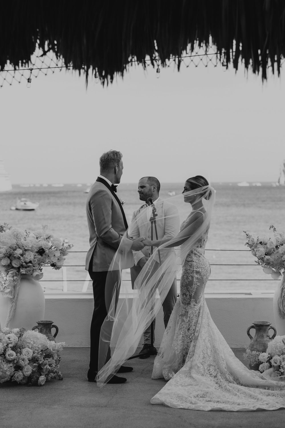 Black and white wedding ceremony photo with floral arrangements and a draped ceiling overlooking the water.