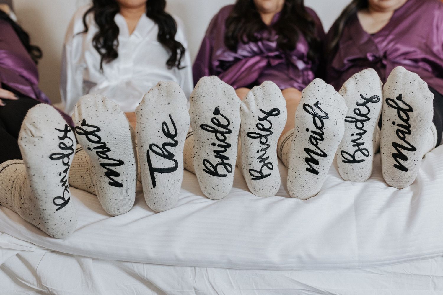 Wedding party members show off their matching gray socks with custom black lettering while sitting together on white bedding.