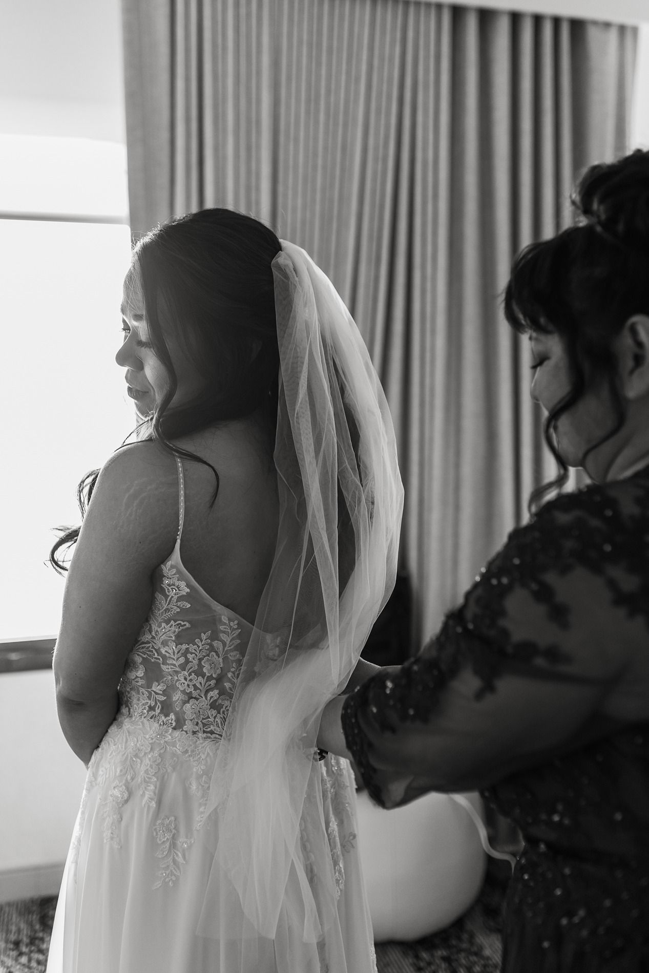 A bride in a white lace wedding dress with a veil stands in front of a window with curtains.