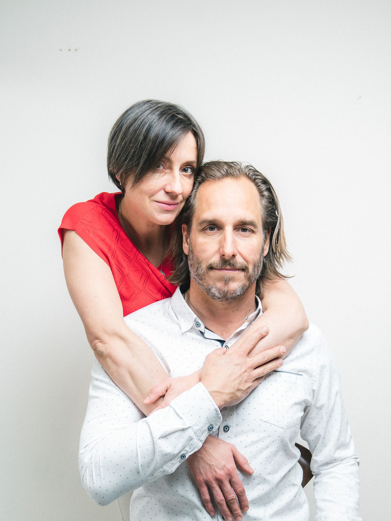 A man and woman pose together smiling, man in dark shirt with arm around woman in white knit sweater.
