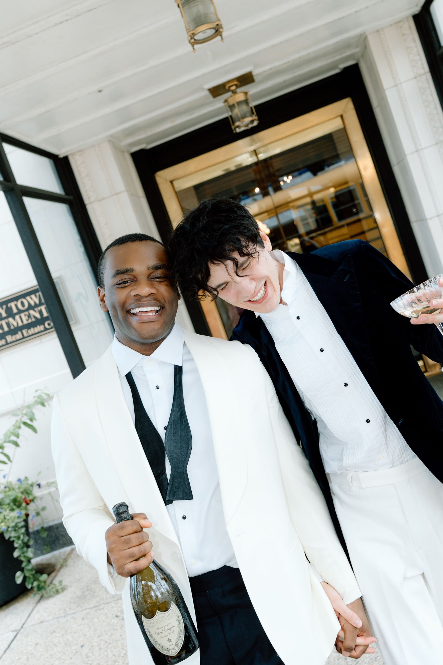 Two grooms in white tuxedos celebrate with champagne outside a building on their wedding day.