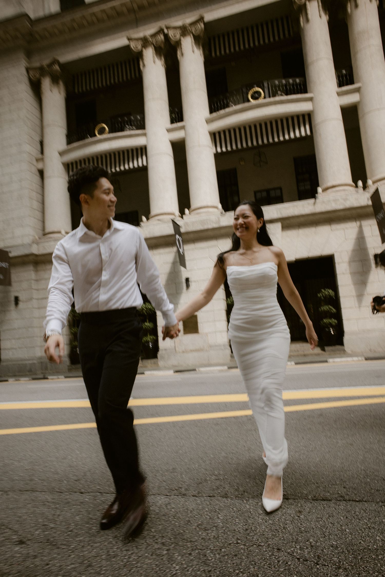 Bride and groom holding hands crossing city street with classical building columns backdrop.