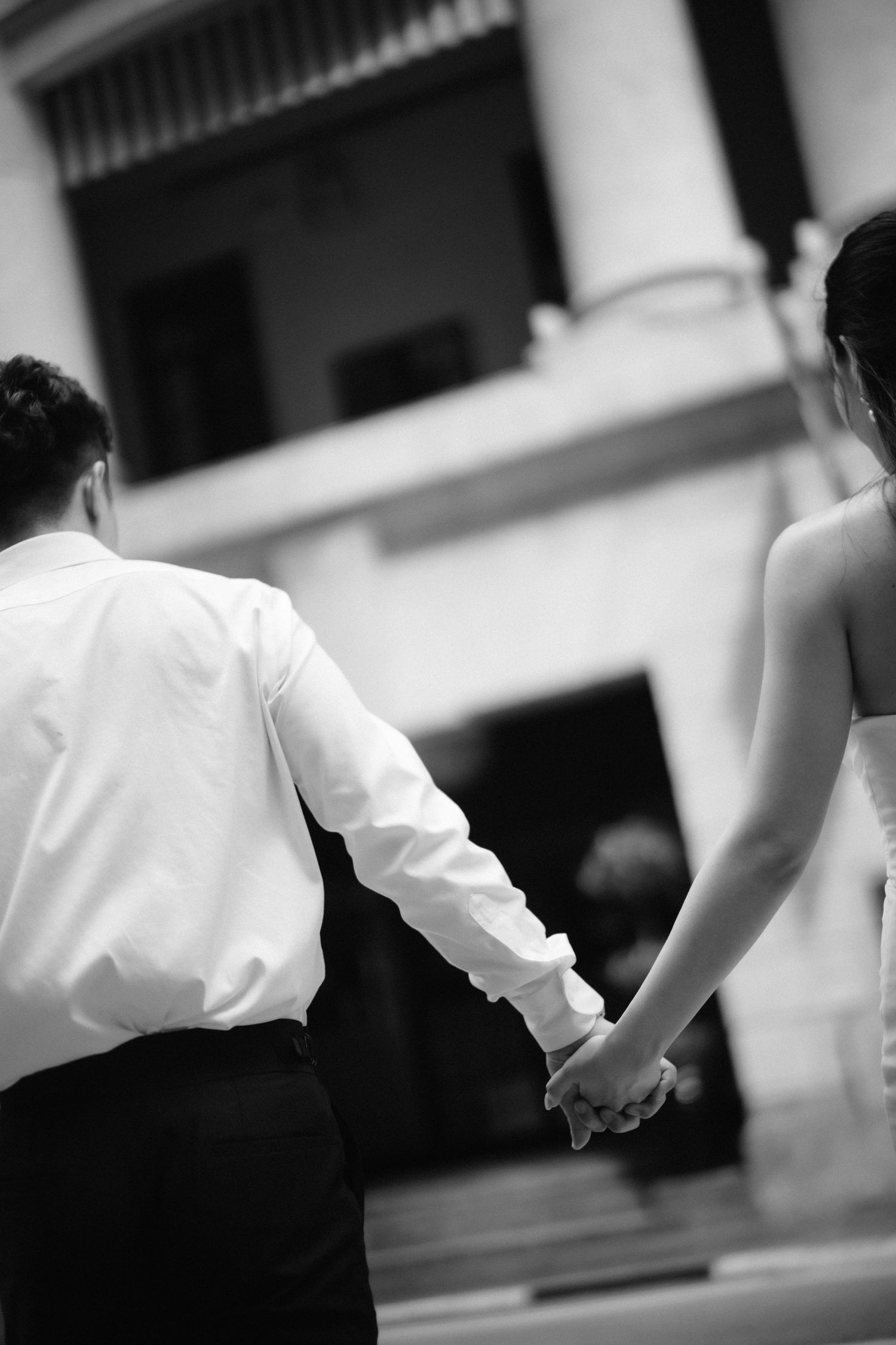 Black and white photo of newlyweds holding hands walking toward classical architecture.