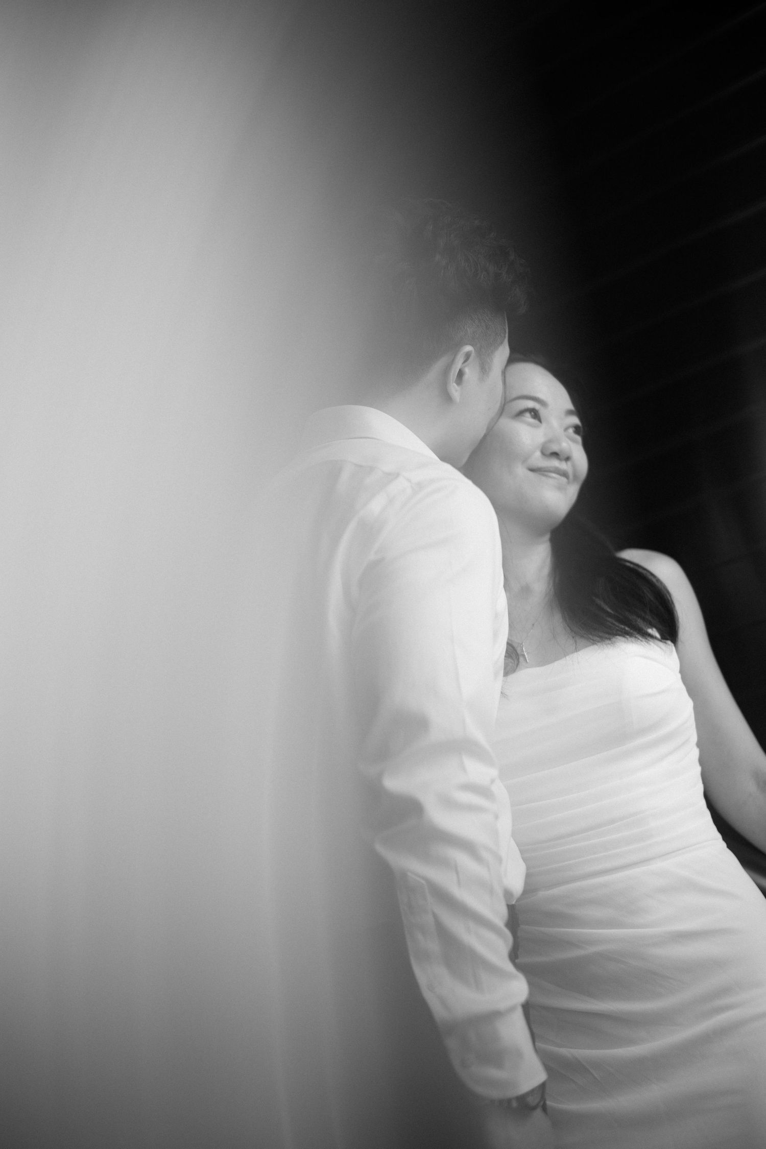 Black and white romantic portrait of bride leaning against groom near light and shadow contrast.