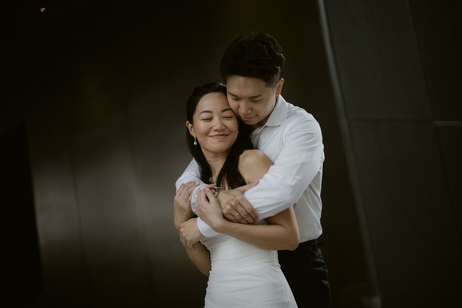 Couple sharing a tender embrace, woman in white dress smiling with eyes closed, man hugging her from behind.