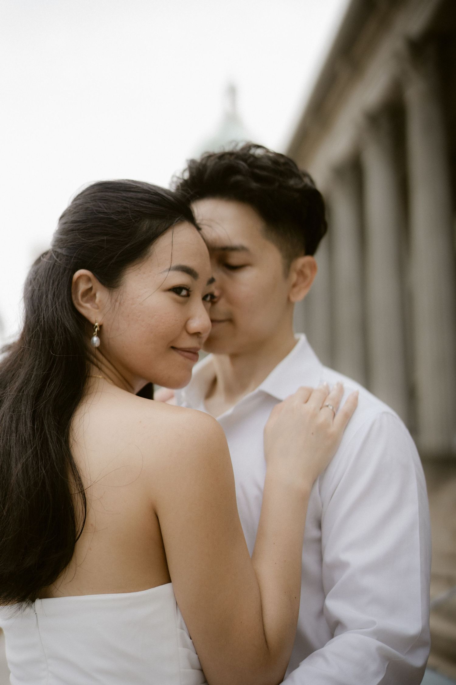 Bride in strapless white dress gazes at camera as groom kisses her temple near classical stone columns.