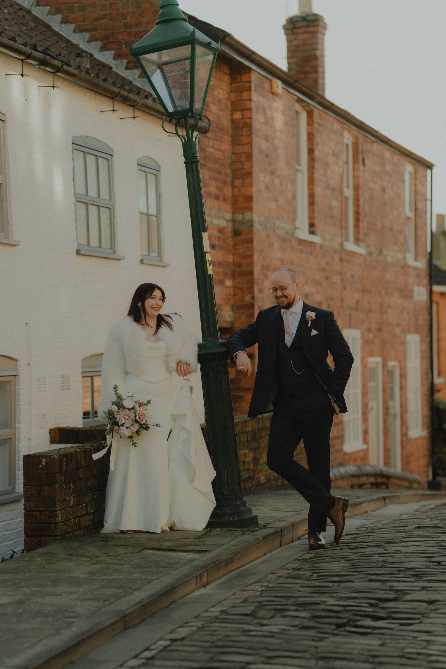 Bride and groom pose by a vintage green lamppost on cobblestone street, surrounded by charming brick buildings.