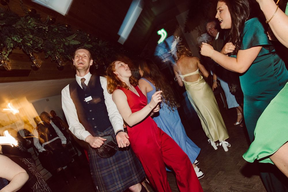 Party guests dancing and celebrating at a festive event with someone wearing a traditional Scottish kilt.