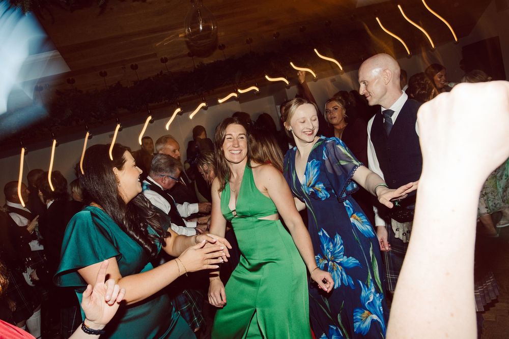 Wedding guests in green and blue attire dance enthusiastically on a dimly lit dance floor.