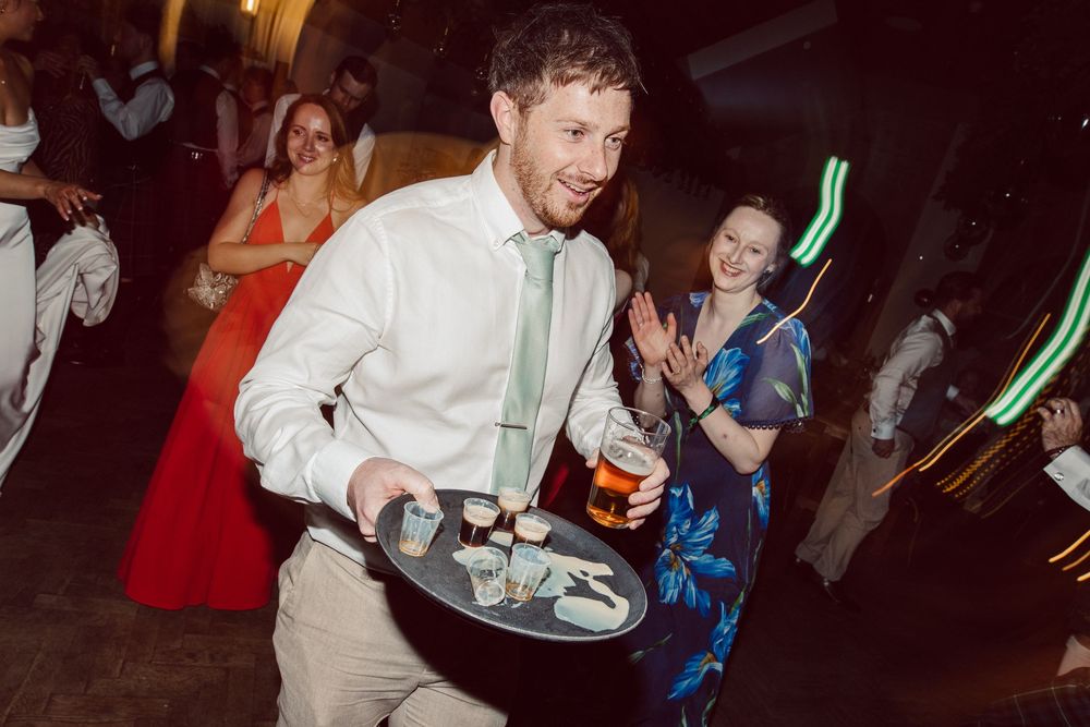 People dancing and celebrating at a wedding reception with glowing green light sticks on a dark dance floor.