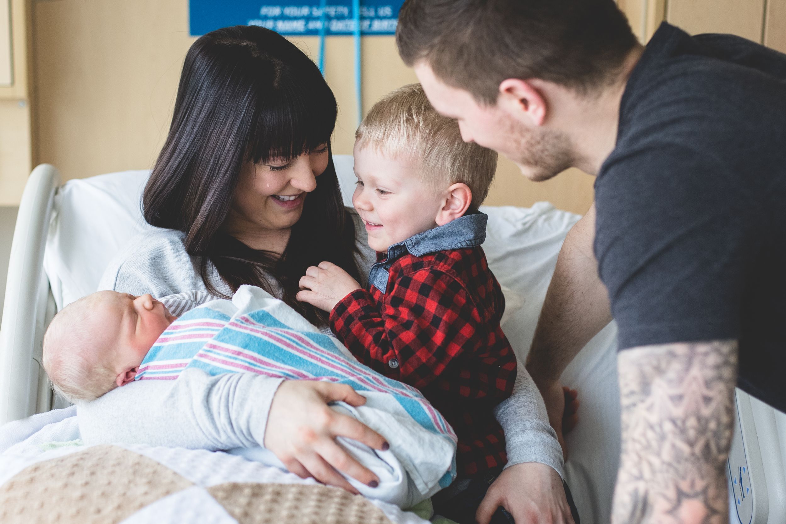 Big brother is meeting newborn baby brother at Montfort hospital. During Fresh 48 photo session. Mom is smiling and has a tear on her cheek.