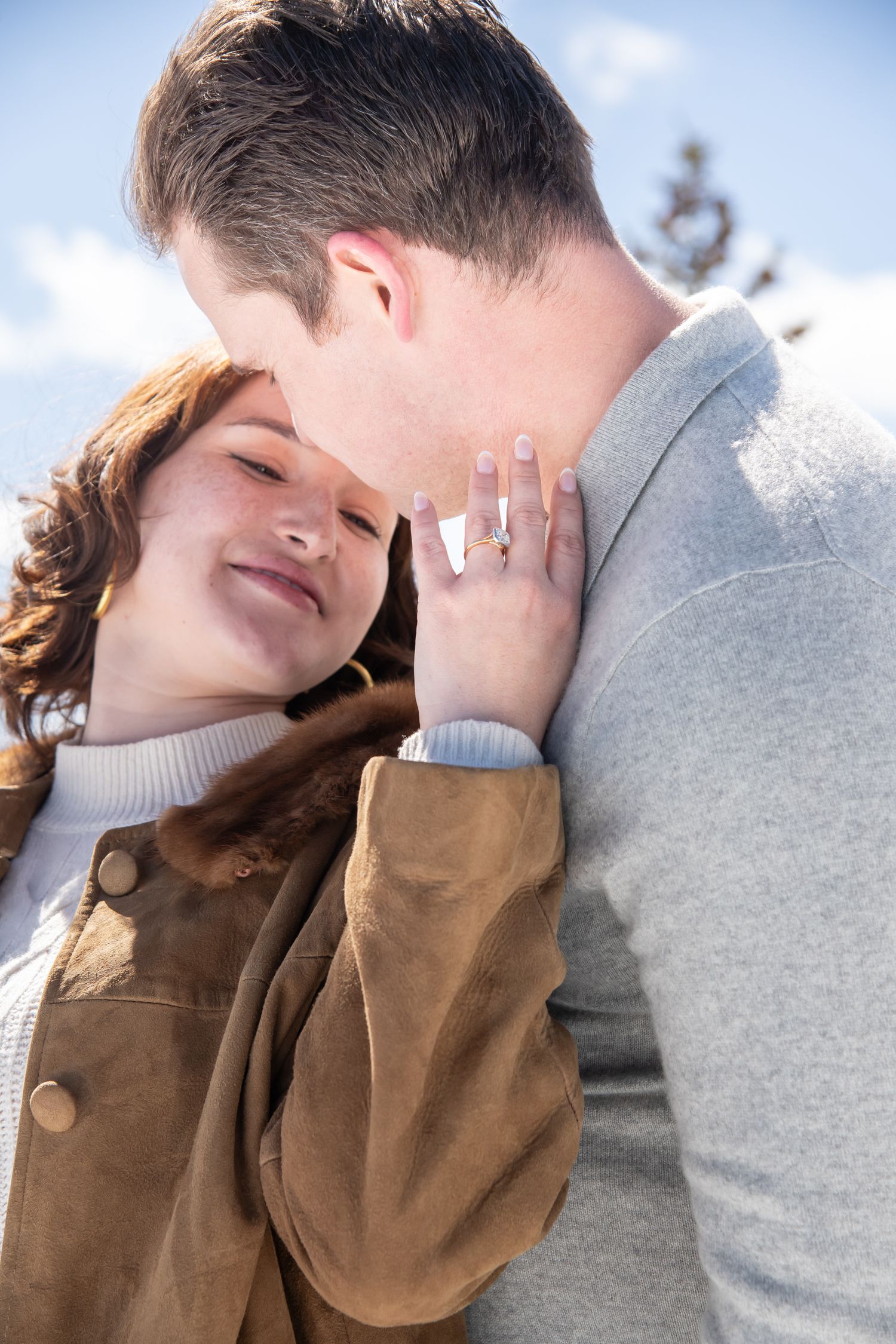 A couple in casual winter attire share an intimate moment against a bright sky background during Aspen winter proposal photos.
