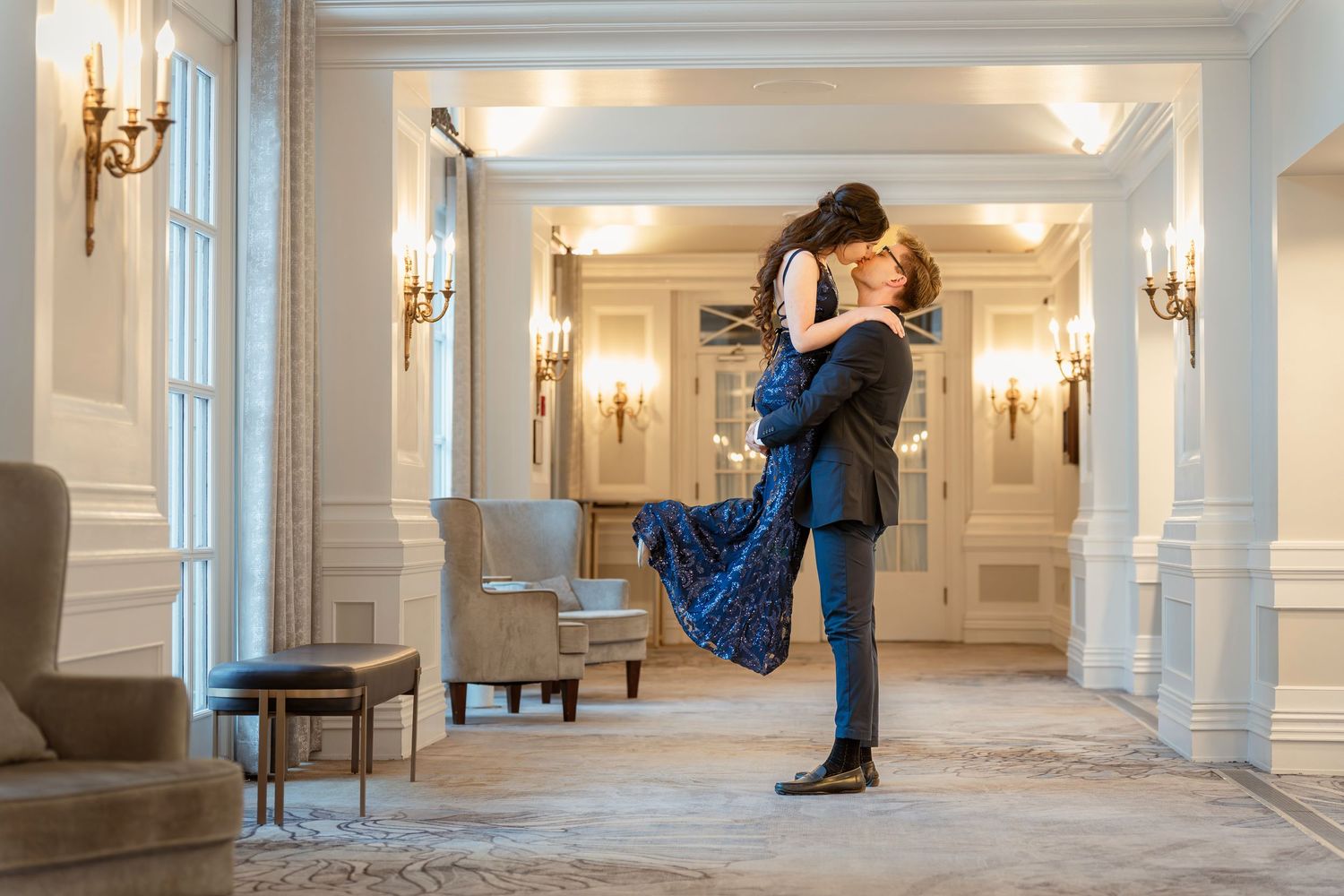 A couple dancing in an elegant white ballroom with ornate wall sconces and decorative moldings.