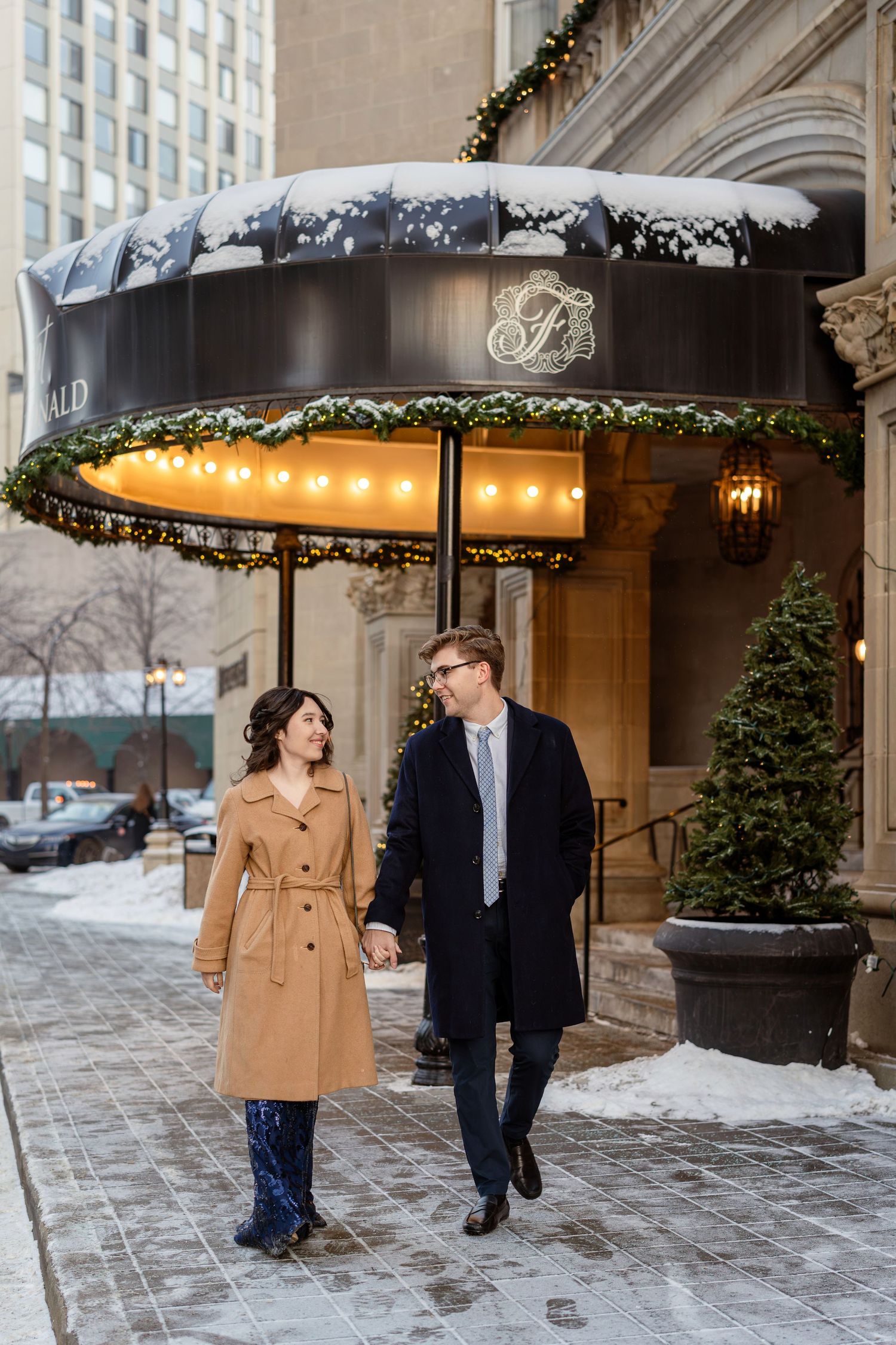A couple walks hand in hand outside a luxury hotel entrance with winter decor and snow on the ground.