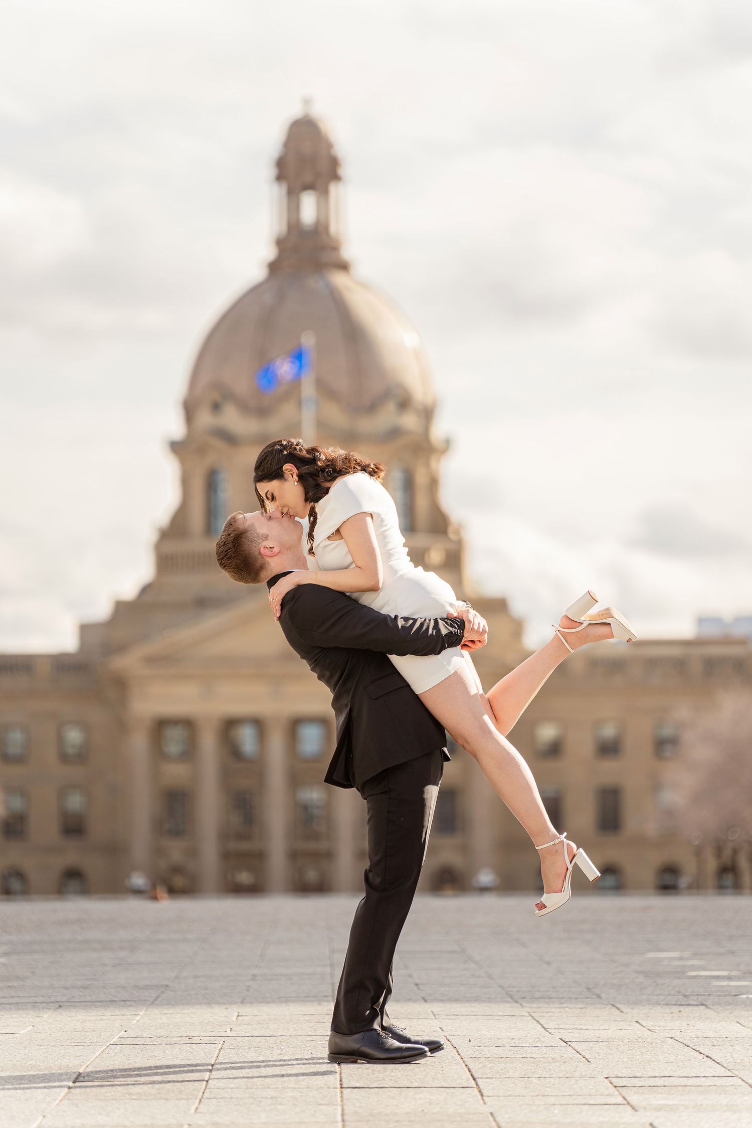 Groom lifting bride in front of Alberta Legislature Building dome during outdoor wedding photos.