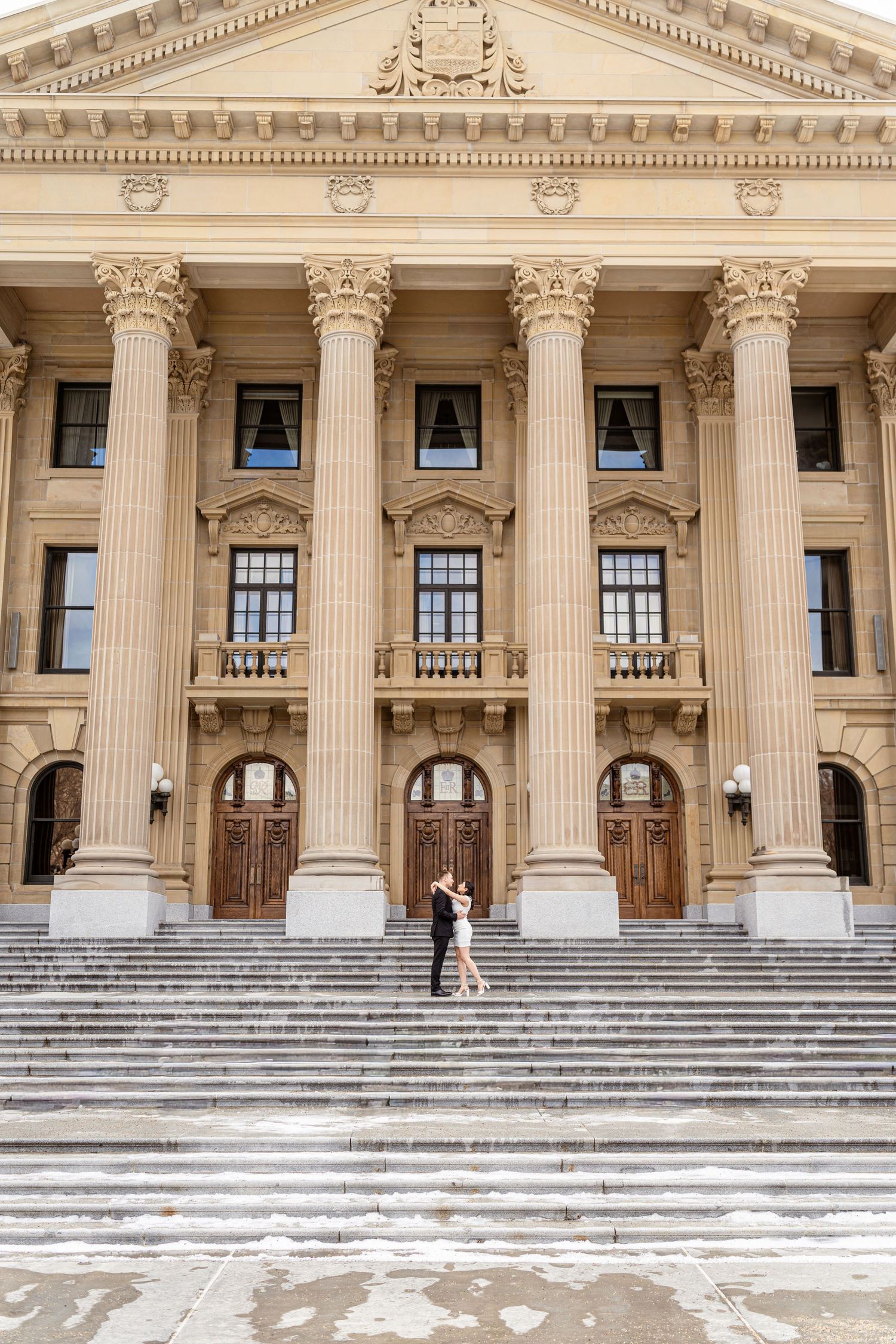 Couple embracing on steps of grand neoclassical building with tall Corinthian columns and ornate facade.