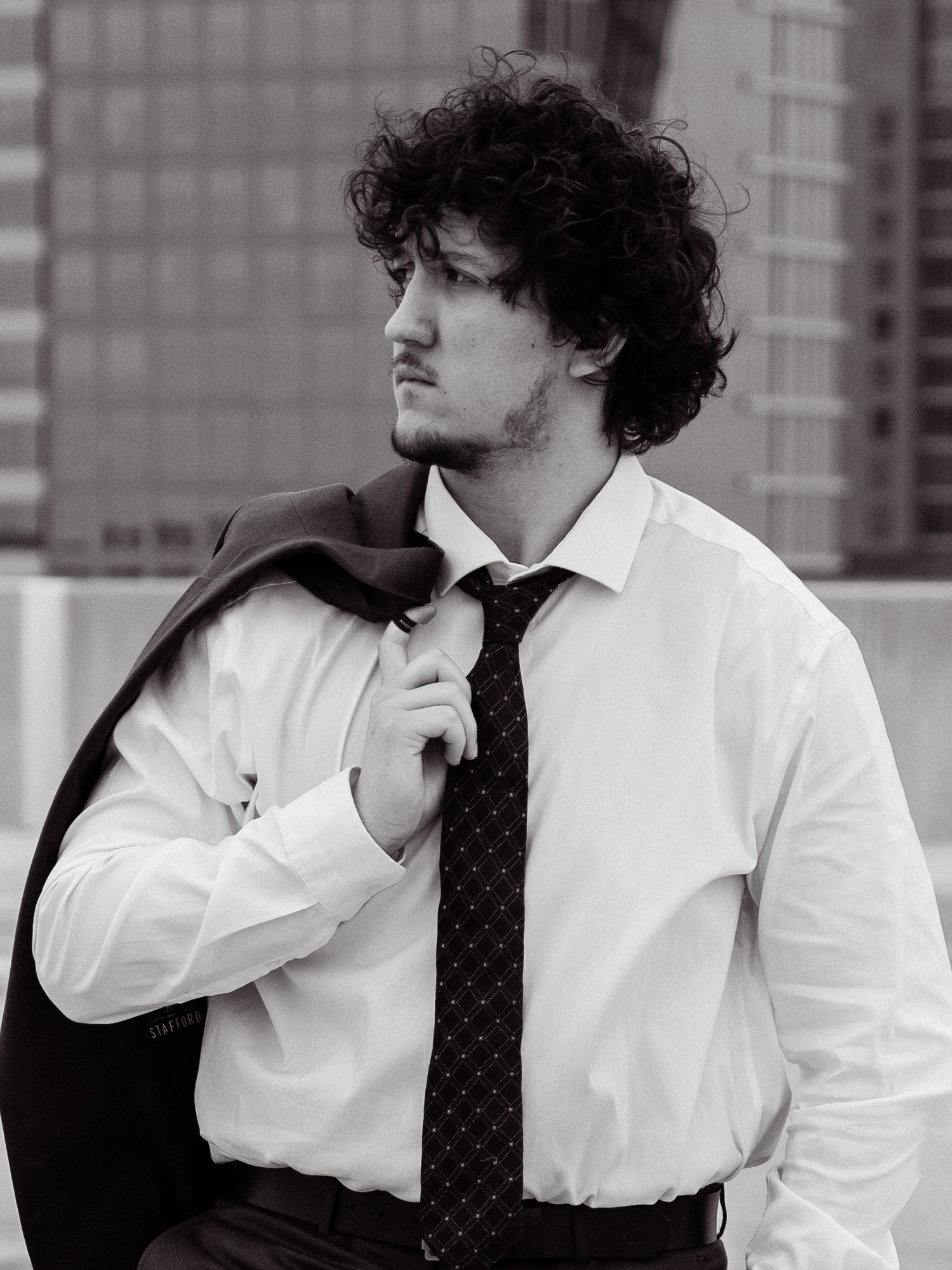 Black and white portrait of a person in business attire with curly hair and tie looking away from camera against urban backdrop.