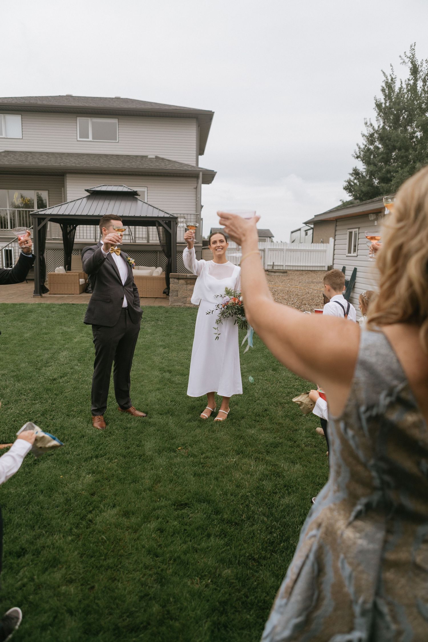 Wedding celebration on backyard lawn with guests toasting under cloudy sky.