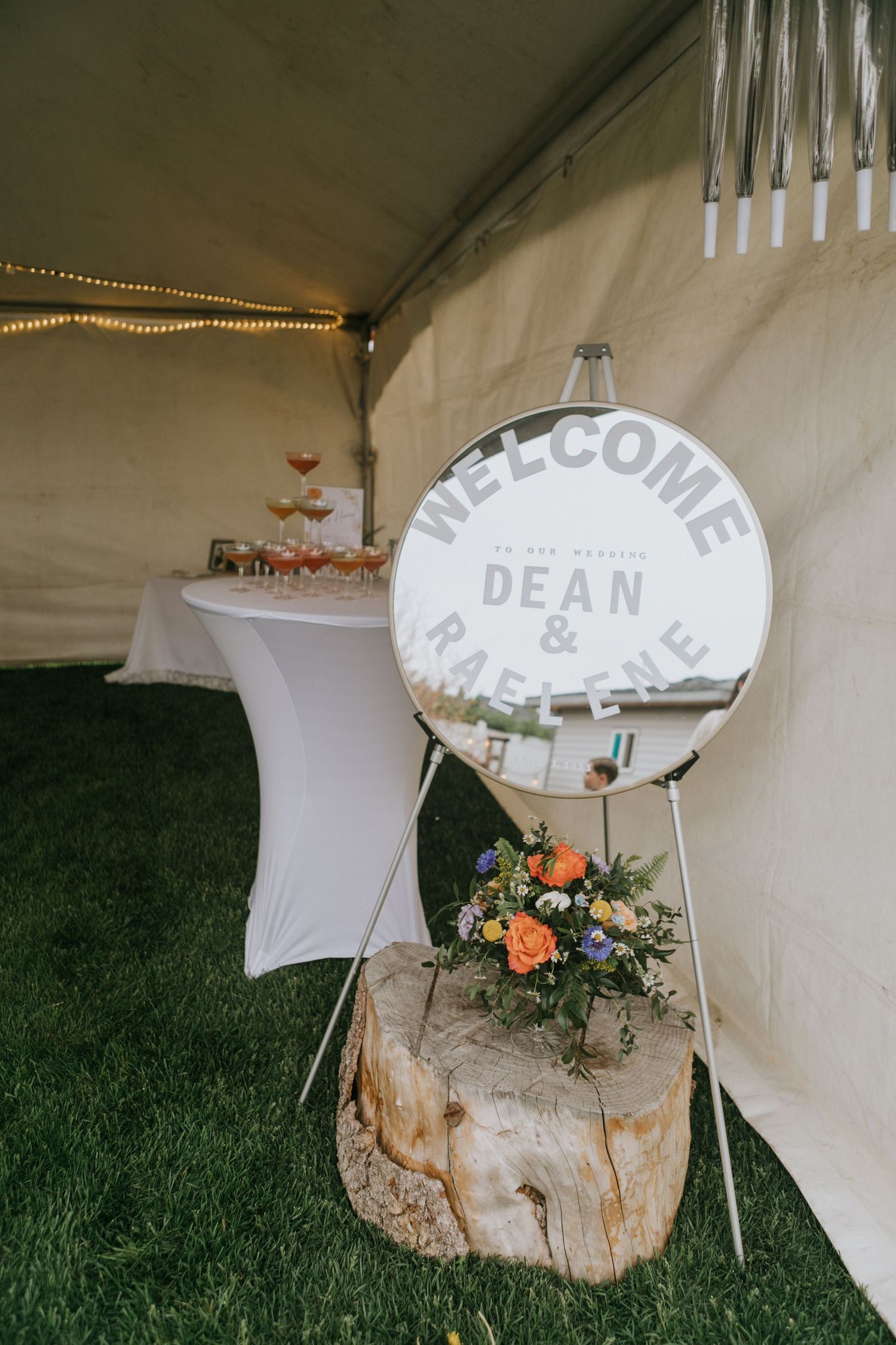 Welcome sign and floral arrangement displayed on rustic wood stump at outdoor wedding reception.