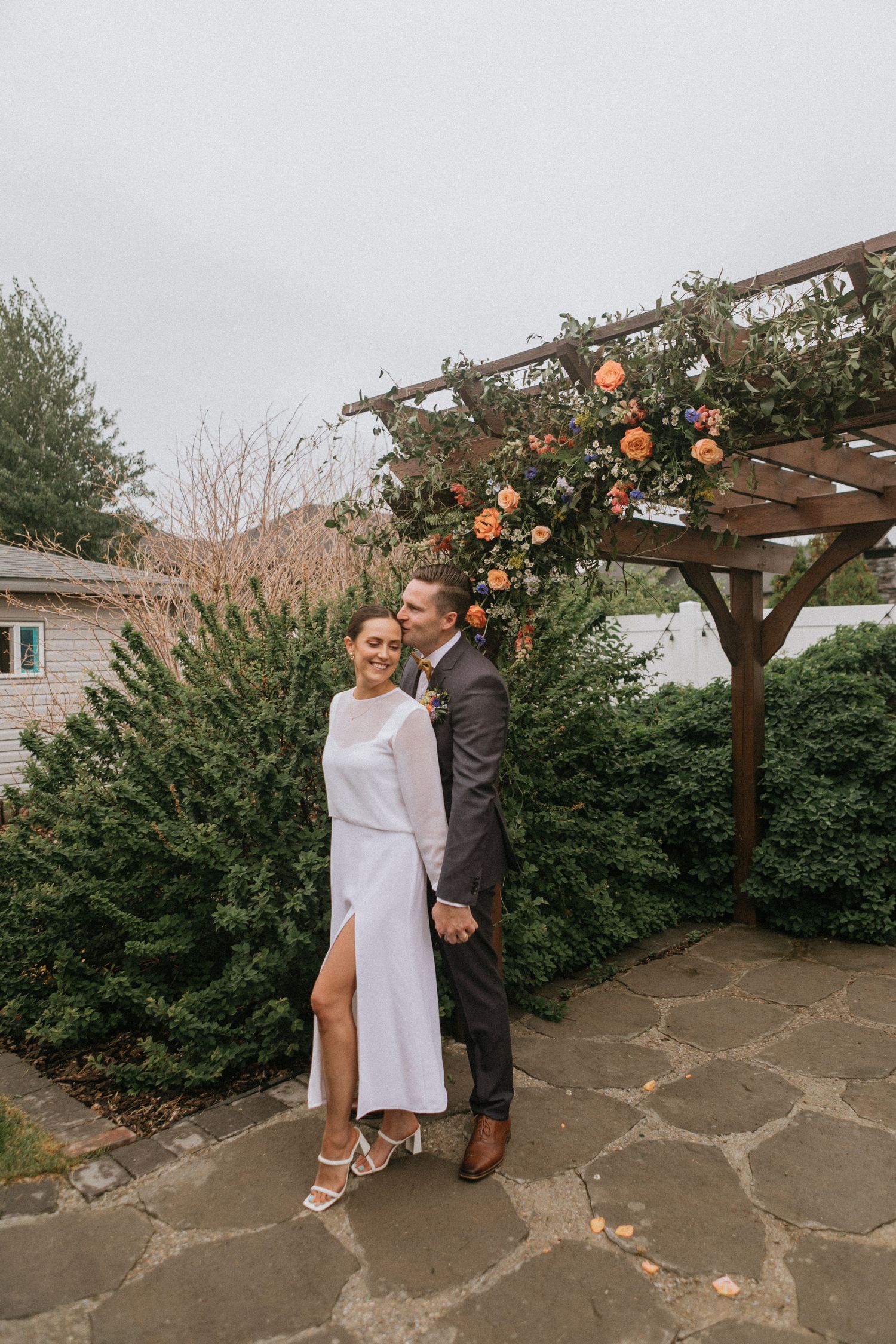 Elegant couple posing under wooden pergola adorned with peach-colored climbing roses.