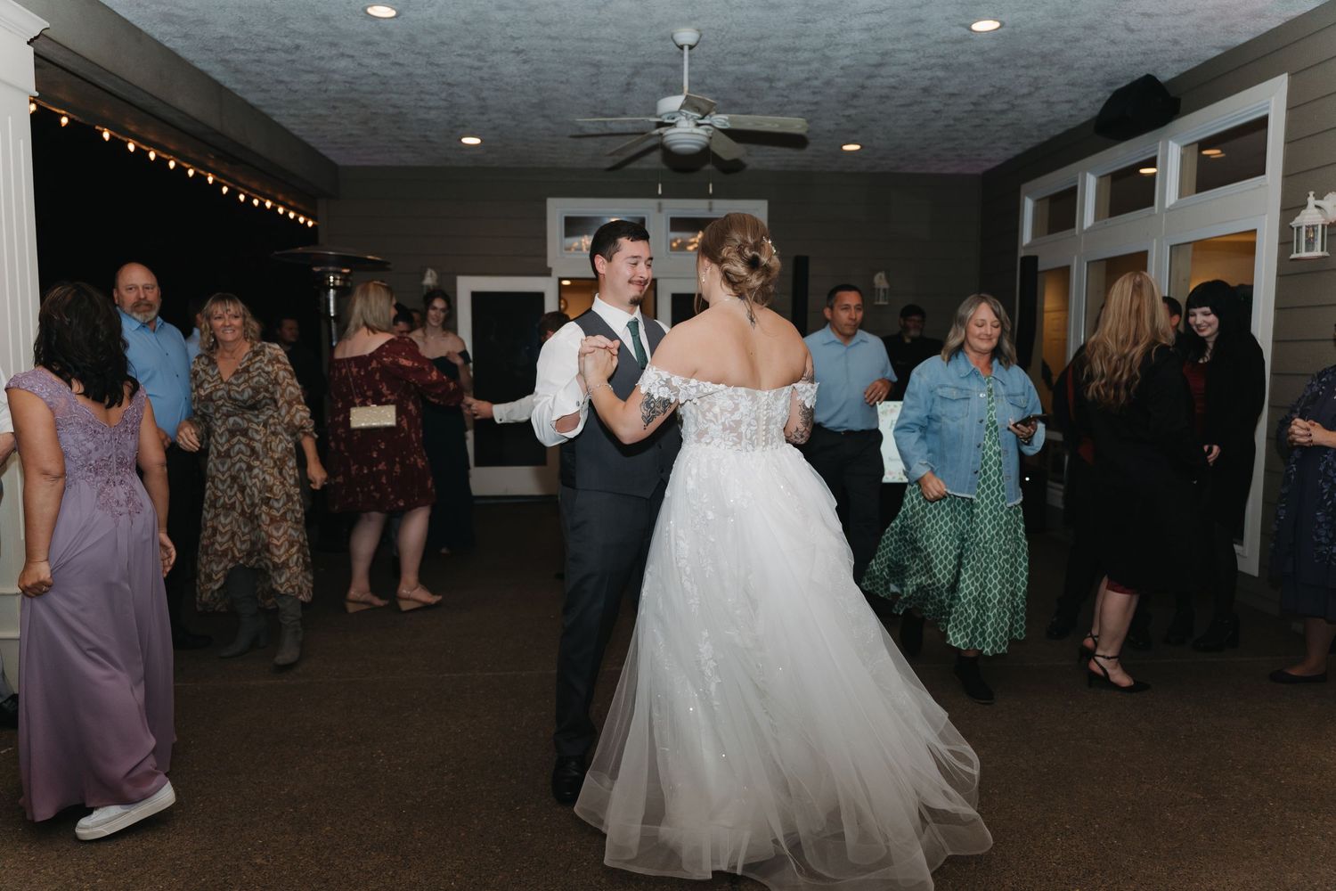 People dance and socialize at a wedding reception in a venue decorated with string lights and featuring a white gown.