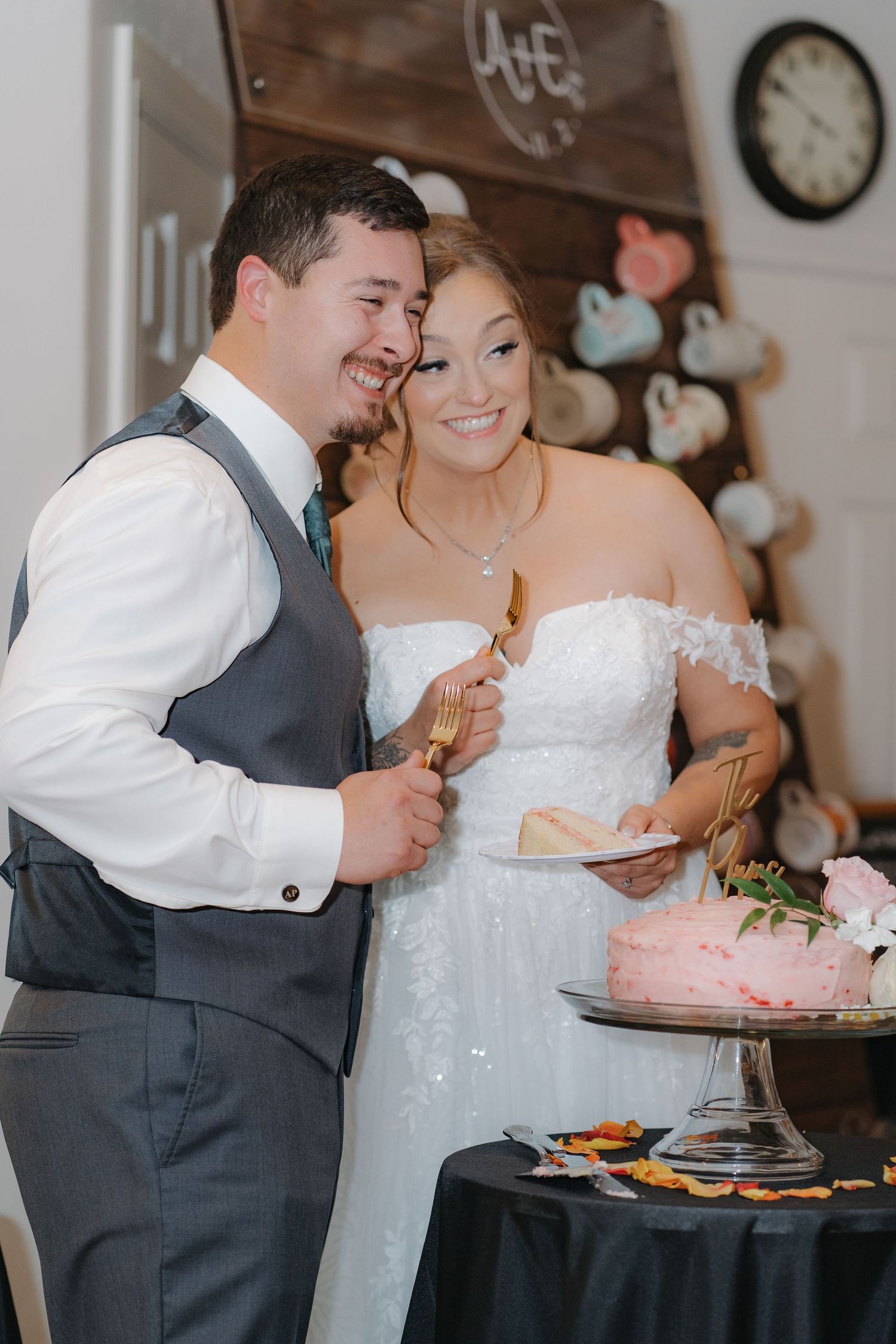 A happy couple sharing a laugh while cutting their pink wedding cake at their reception with heart decorations in background.
