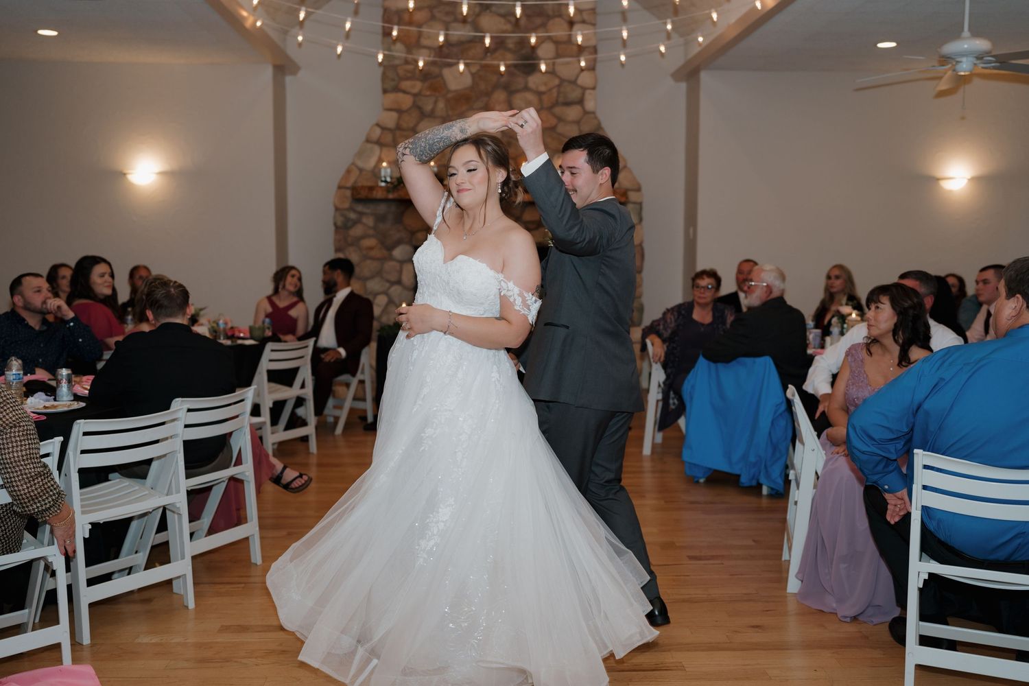 A couple shares a dance in a rustic indoor venue with exposed beams and string lights as guests watch from white chairs.