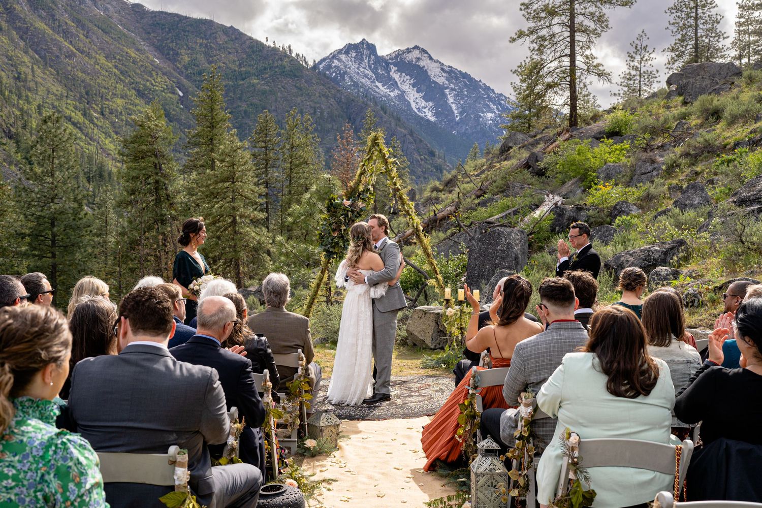 Outdoor wedding ceremony in progress with dramatic mountain landscape and evergreen forest backdrop.