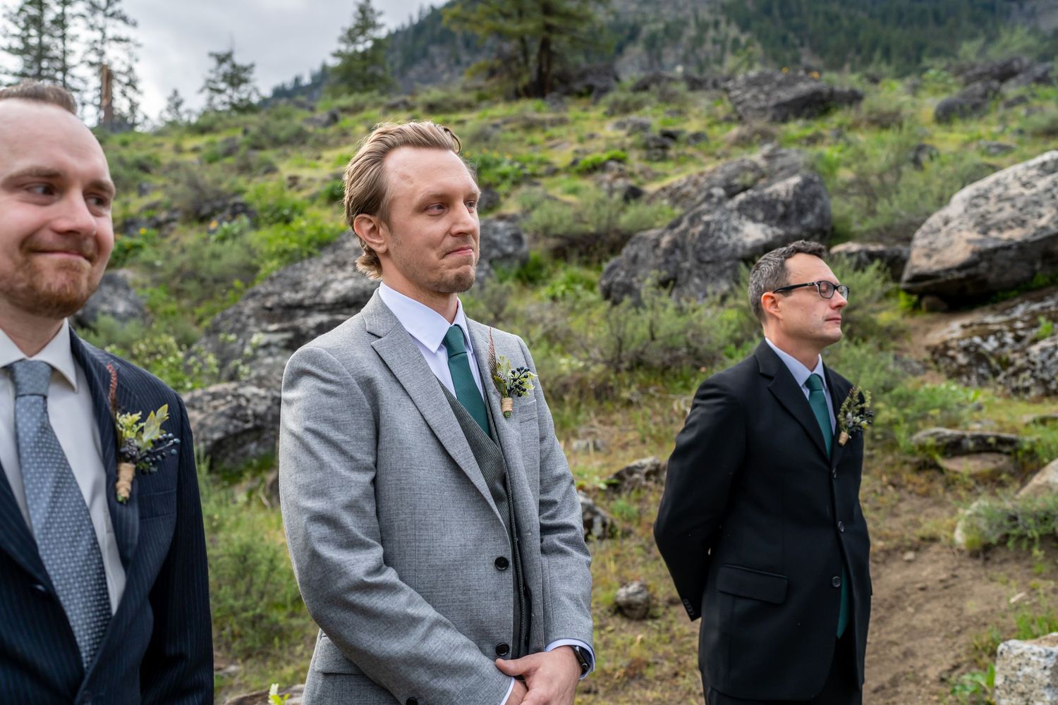 Groomsmen in formal attire stand outdoors against a mountainous backdrop during a wedding ceremony.