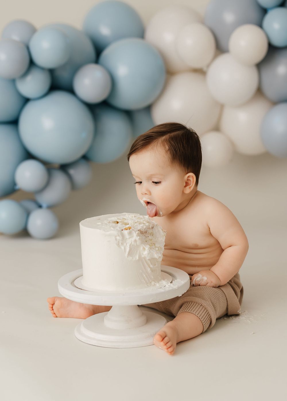 Baby in tan pants exploring white cake with blue and white balloon arch background.