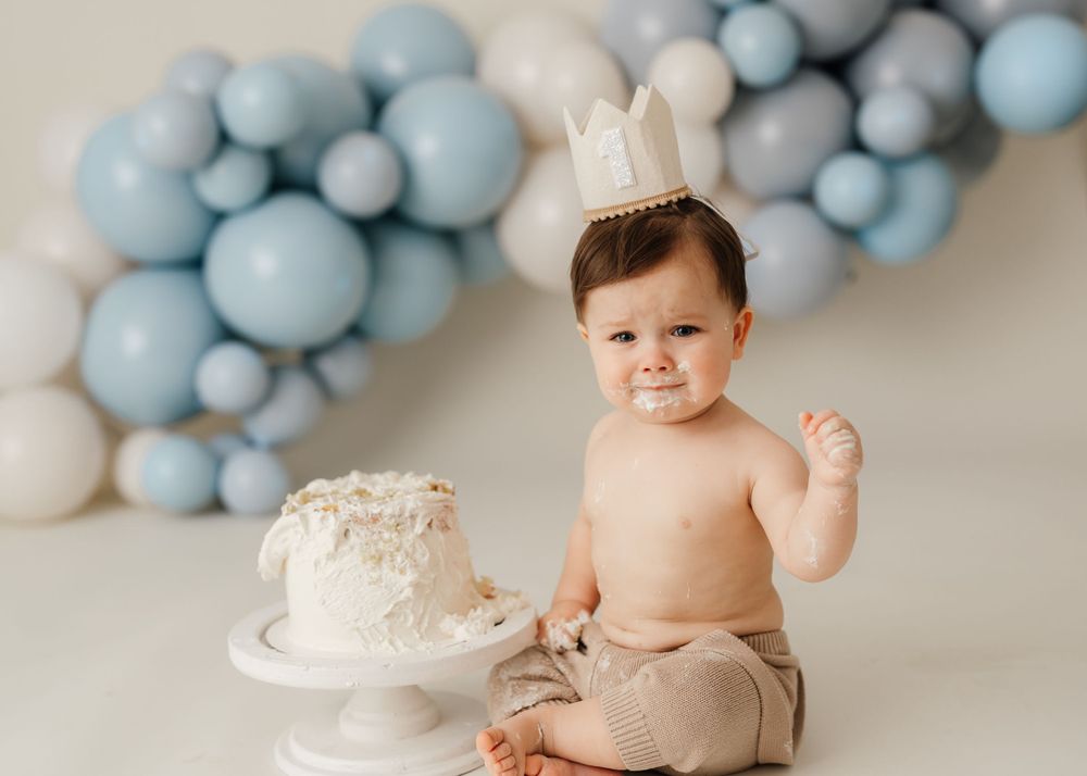 Baby wearing paper crown next to white cake with blue balloon backdrop.