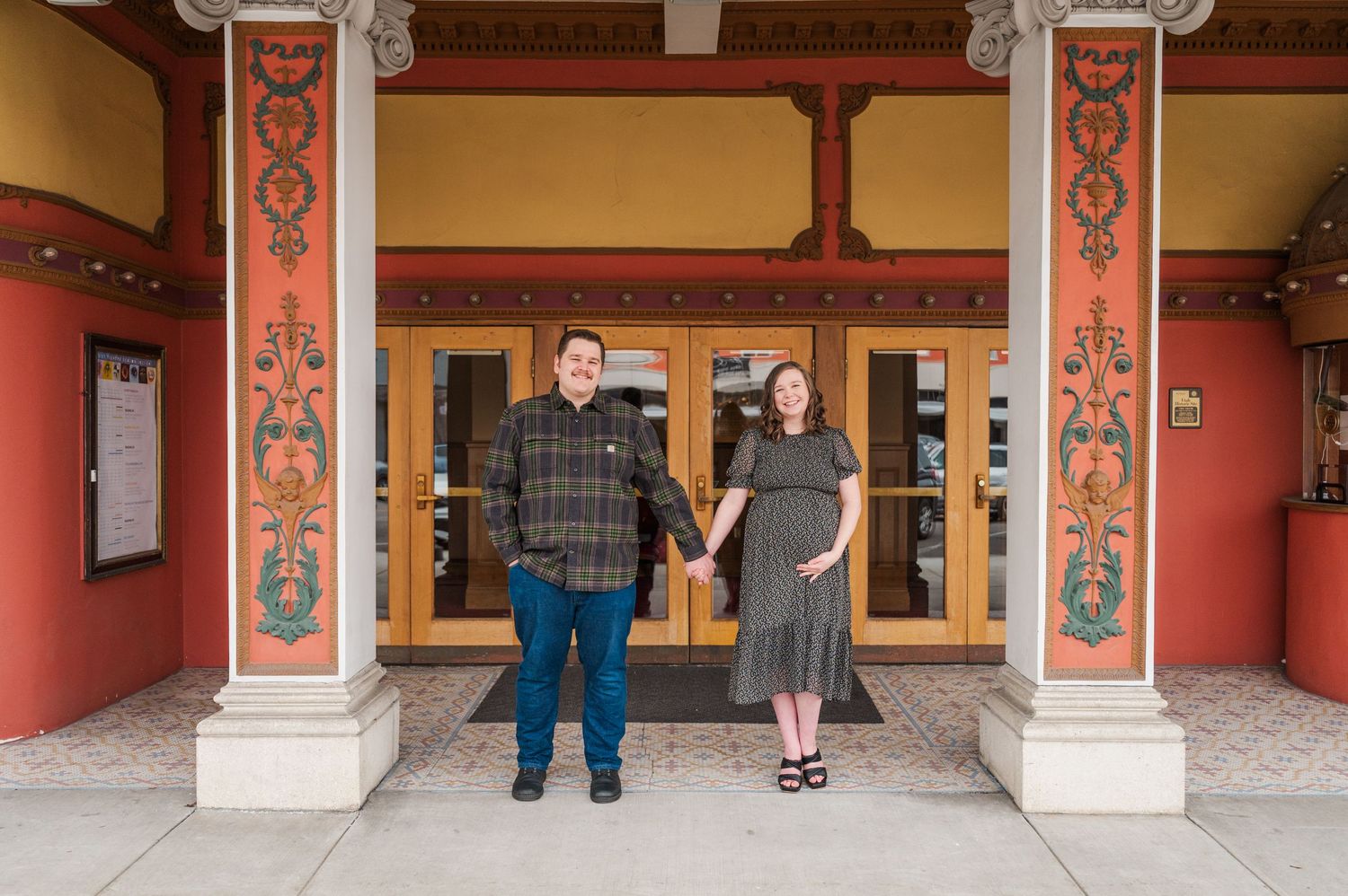 A couple poses under ornate red and orange architectural columns at the entrance of a building.