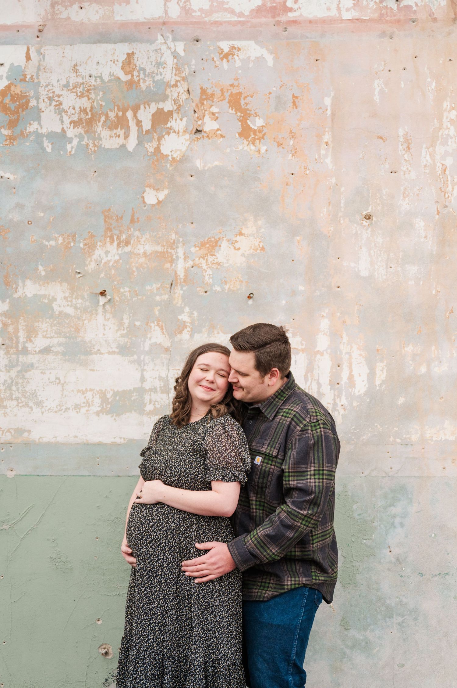A loving maternity photo against a weathered wall with peeling paint textures.