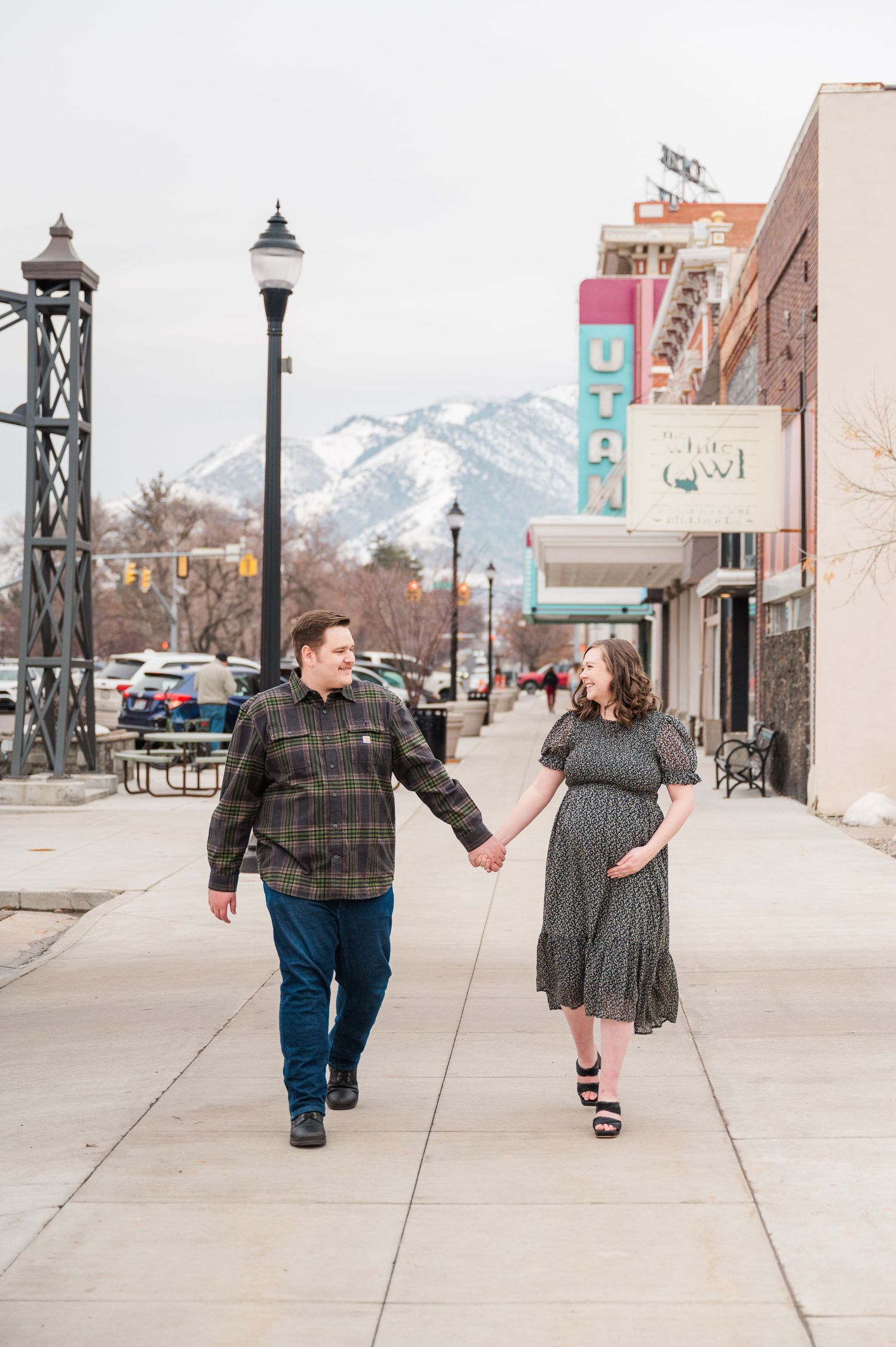 Couple walking hand in hand on sidewalk with snowy mountains visible in background.