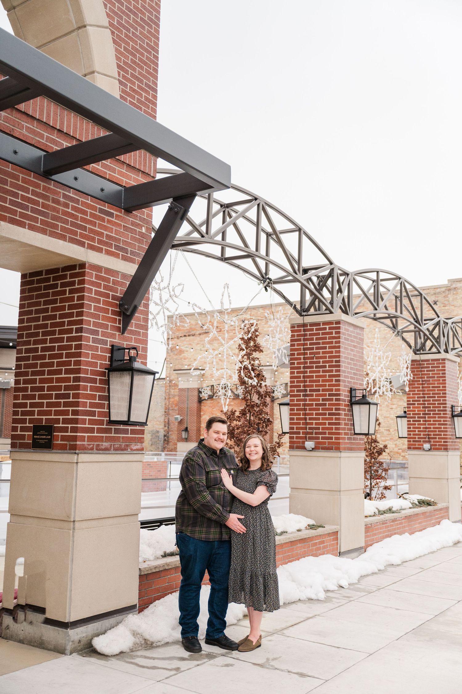 Couple standing beneath metal architectural feature against brick building in winter.