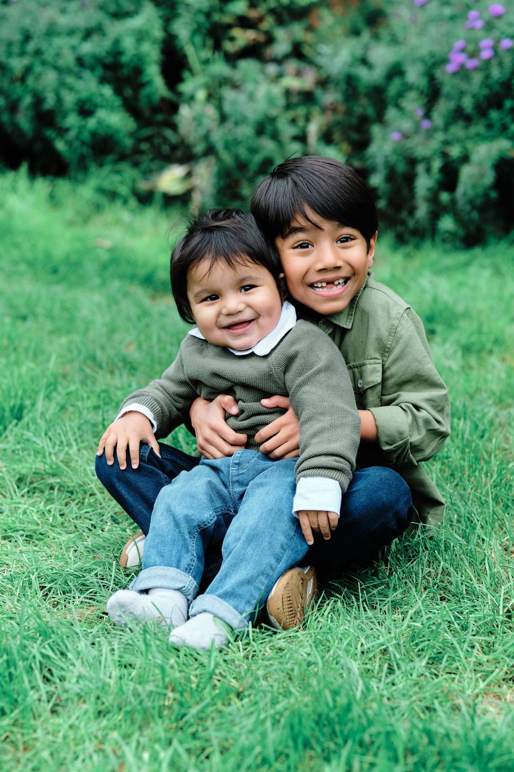 Two children playing and laughing together while sitting on green grass in a garden setting.