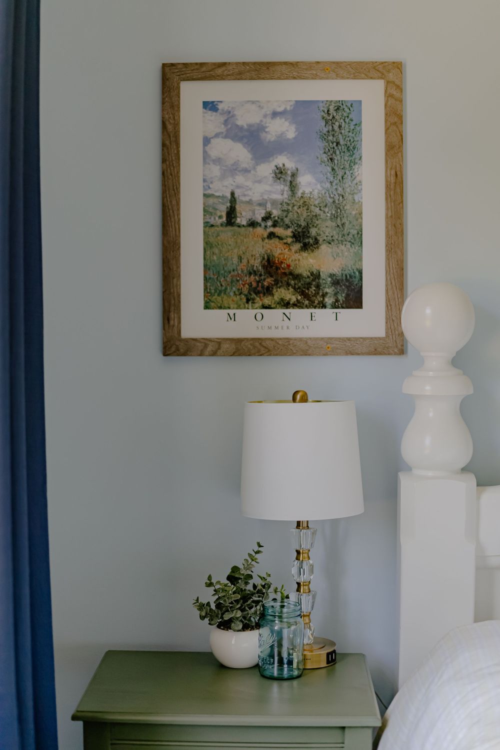 A framed Monet-style landscape print hangs above a green bedside table with white lamp and potted plant in bedroom decor.