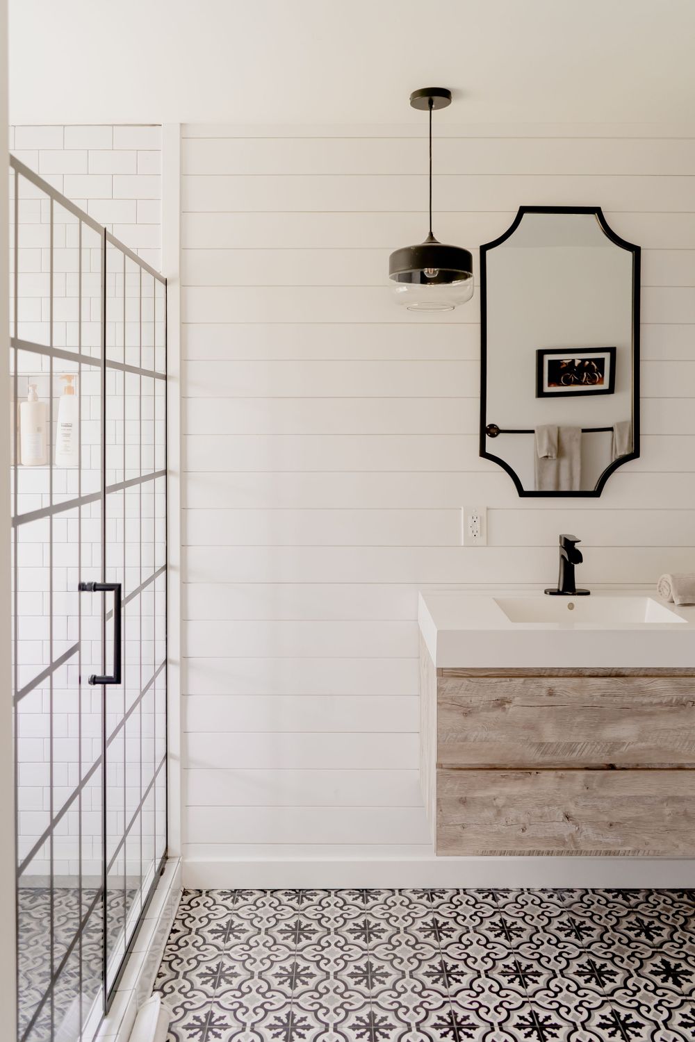 Modern bathroom with patterned floor tiles, white shiplap walls, framed mirror, and glass shower enclosure.