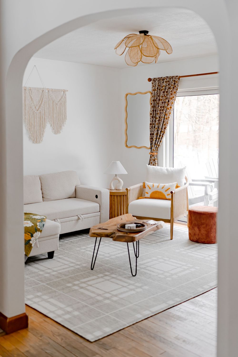 Modern boho living room features white couch, rattan decor, woven wall art and rust pouf under arched doorway opening.