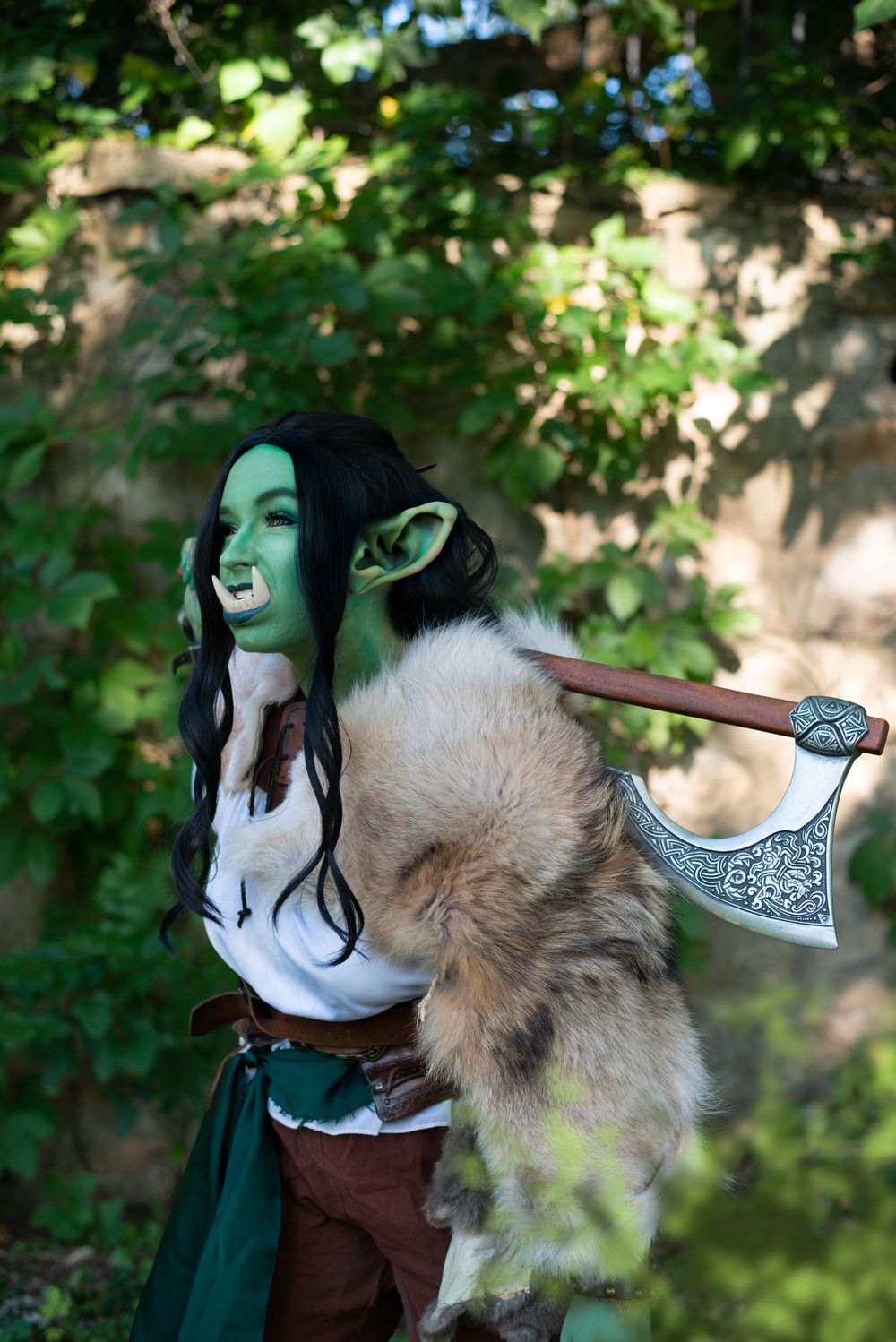 Fantasy cosplayer with green face paint and fur collar holds decorated battle axe in woodland setting.
